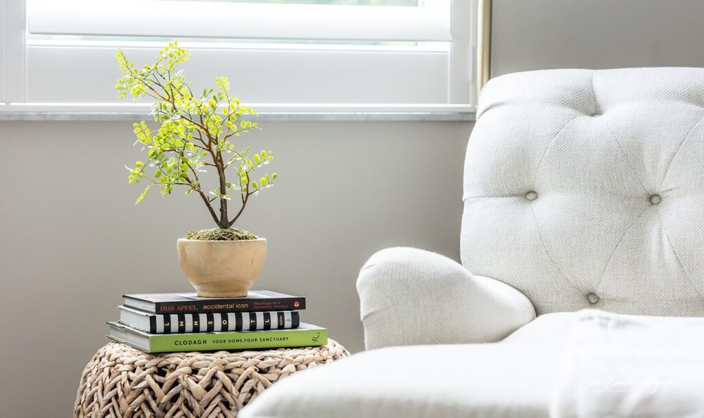 A stack of books placed under a small plant set next to a white love seat